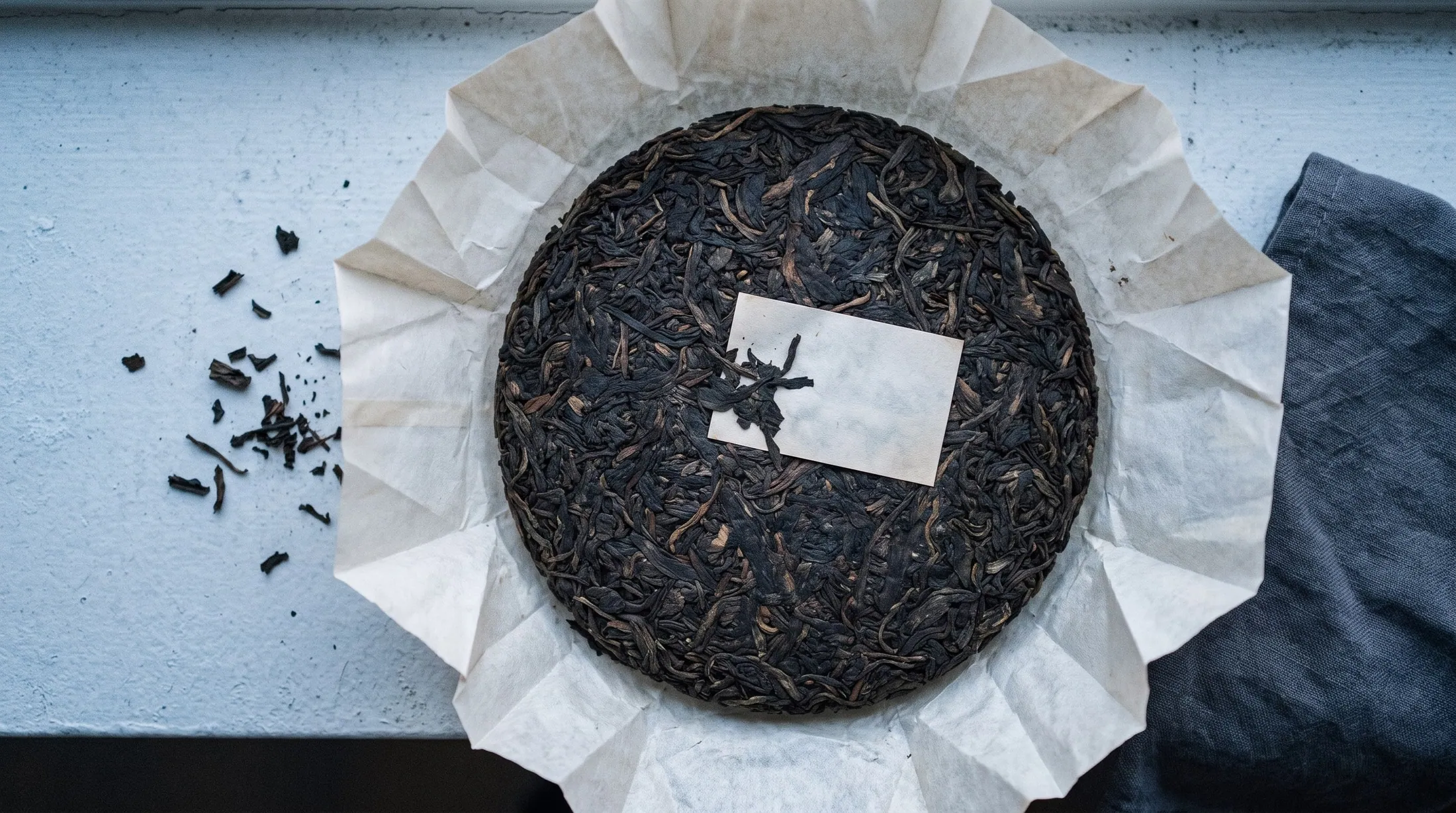 A pu-erh cake in its paper wrapper on a black slate tray, the wrapper partly folded back to show the pressed leaf.