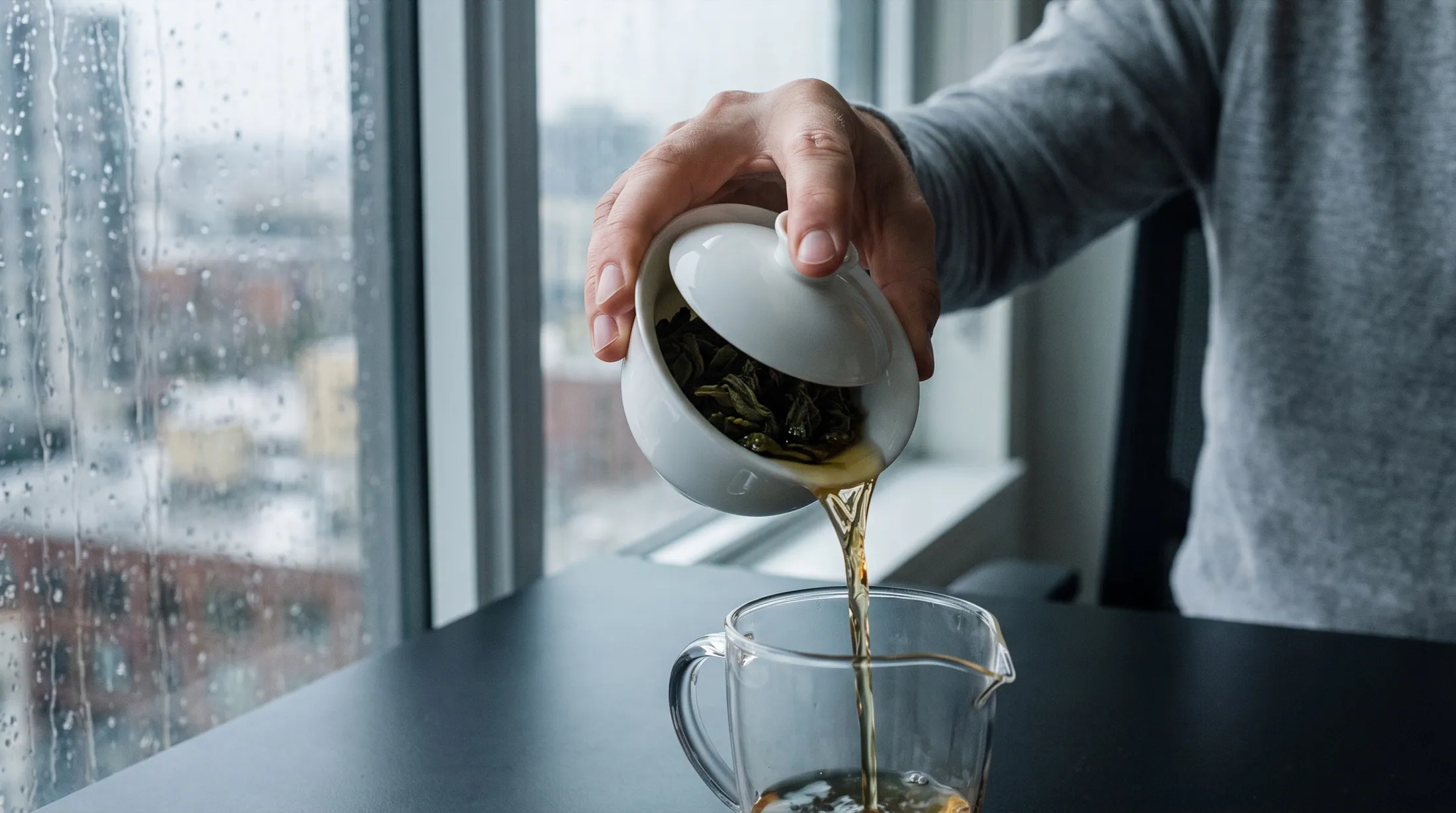 A hand pouring golden oolong from a white gaiwan into a glass pitcher by a rainy window.