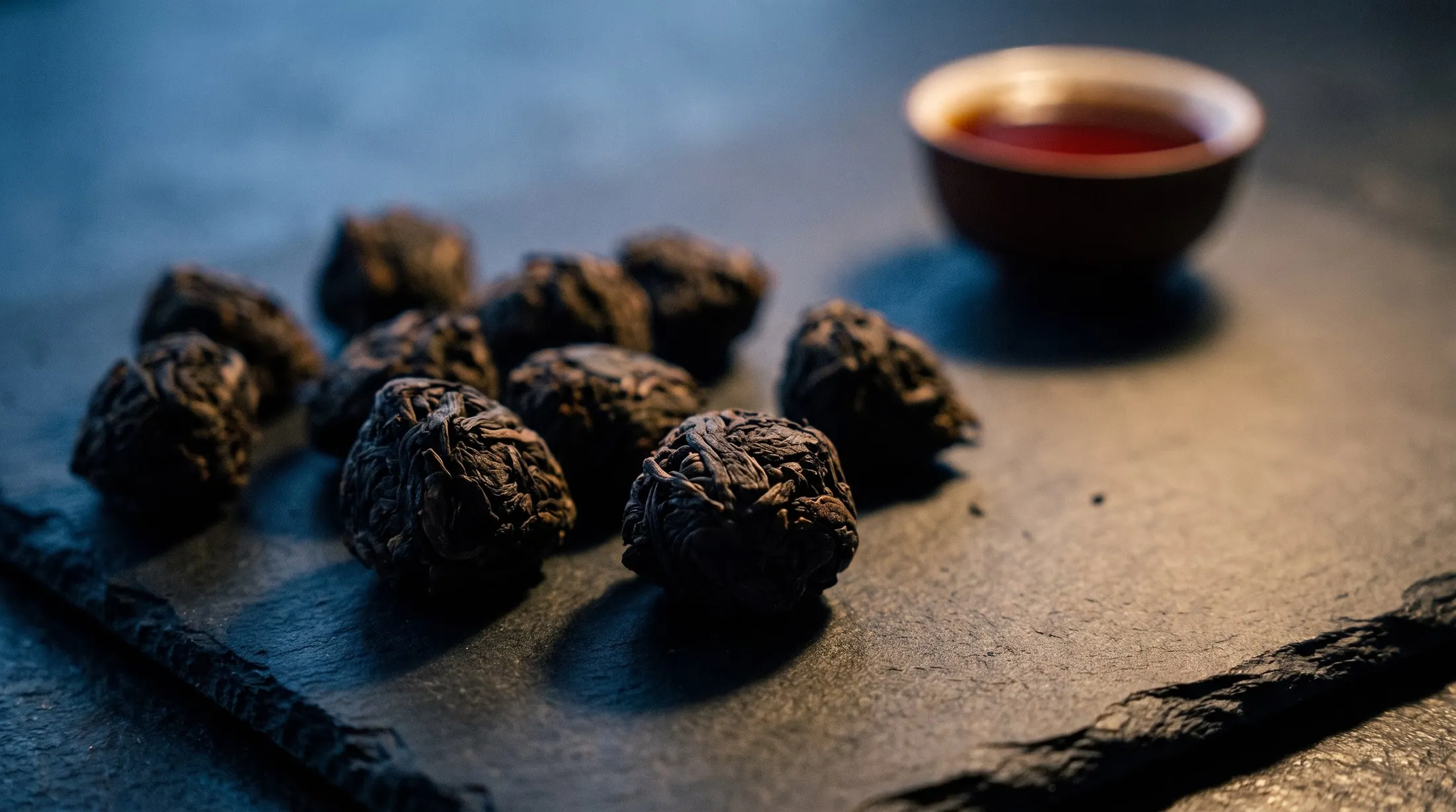 Dark nuggets of Lao Cha Tou shou pu-erh in a black ceramic dish, with a cup of deep red tea beside it.